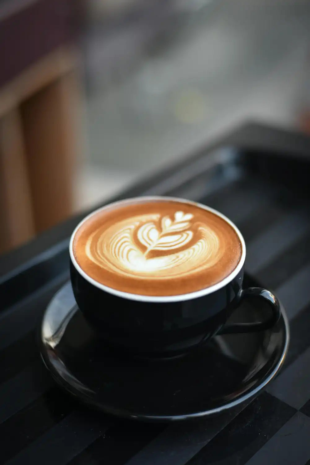 Cappuccino with latte art on a black cup and saucer on a reflective surface.