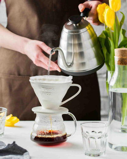 Person making coffee using a pour-over method with a kettle and flowers in the background.